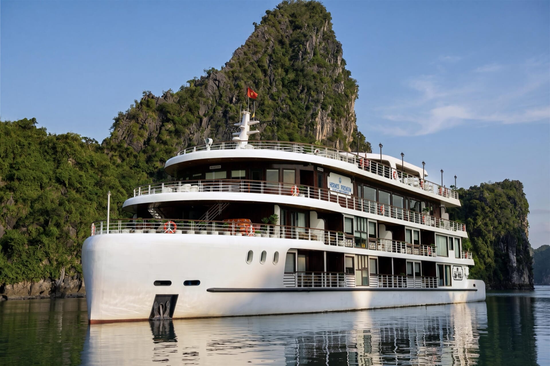 Hermes Premium cruise ship with white hull and wooden terraces in front of a forested limestone island